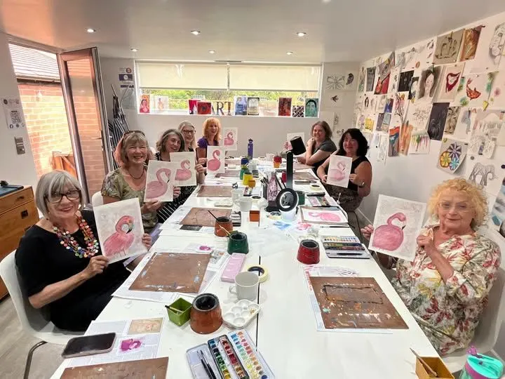A group of adults seated at a long table during an art class, holding up pink flamingo paintings they've created.