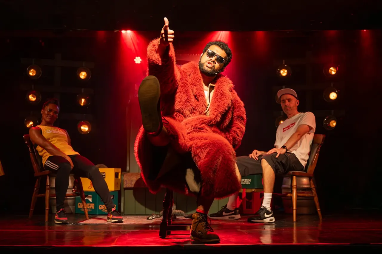 Performer in bright red fuzzy coat and sunglasses strikes a dramatic pose on stage whilst two seated men watch, lit by red stage lights.