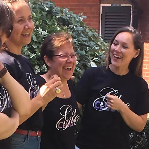 Three women in matching black t-shirts laugh together against a brick wall with climbing ivy.