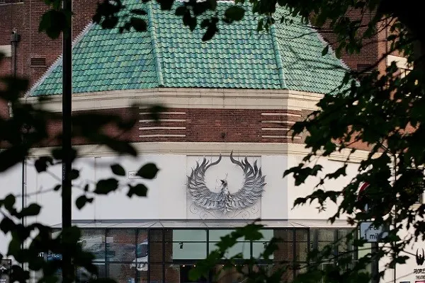 Brick building with turquoise-tiled roof and a large phoenix emblem on its white facade, framed by tree foliage.