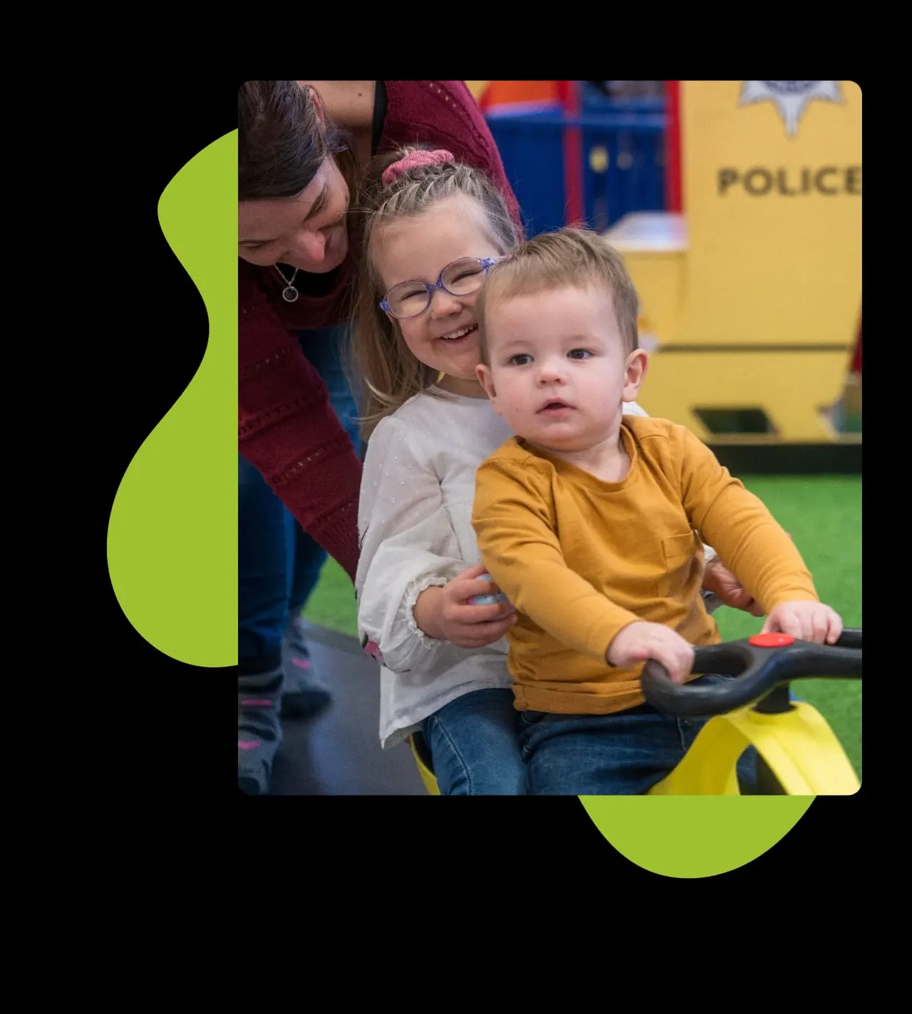 Woman and two children playing on a yellow ride-on toy indoors at a play centre.