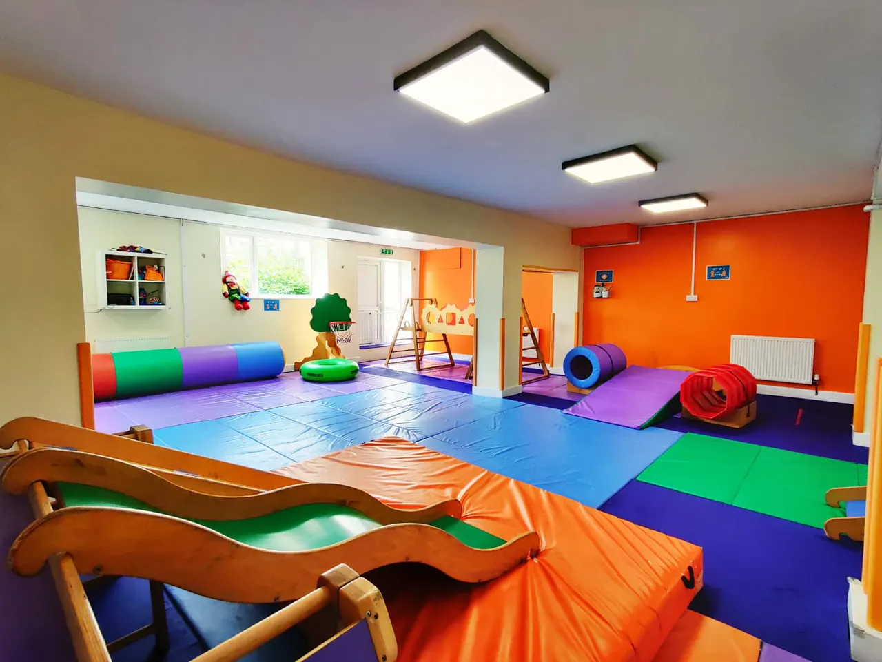 Bright children's soft play area with colourful padded mats, tunnels, climbing equipment and a mirrored wall for activities.