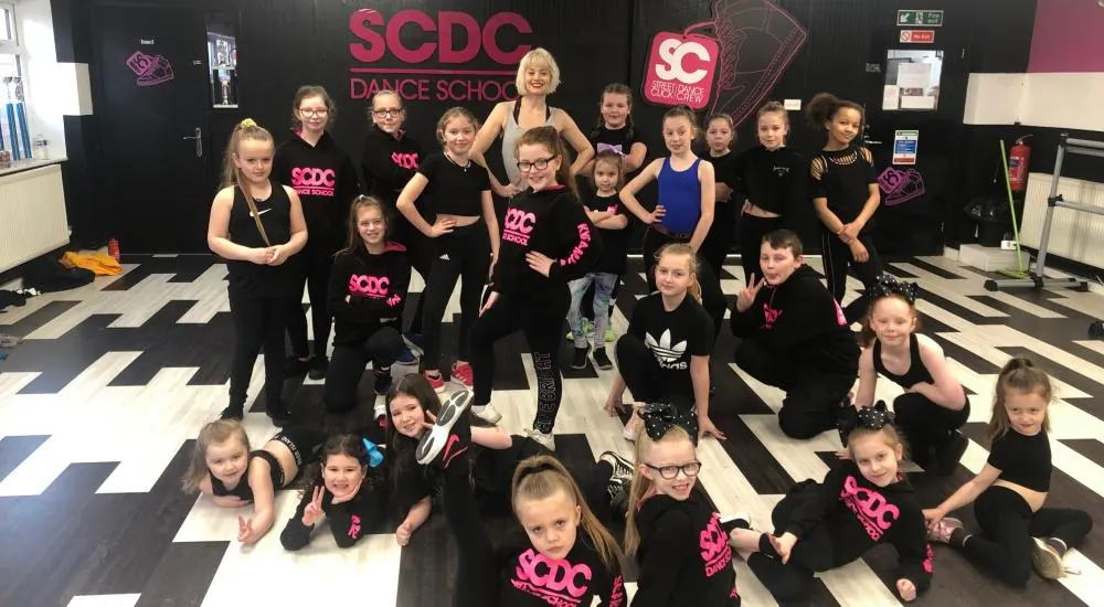 Group of young dancers in black outfits posing with an instructor in a dance studio.