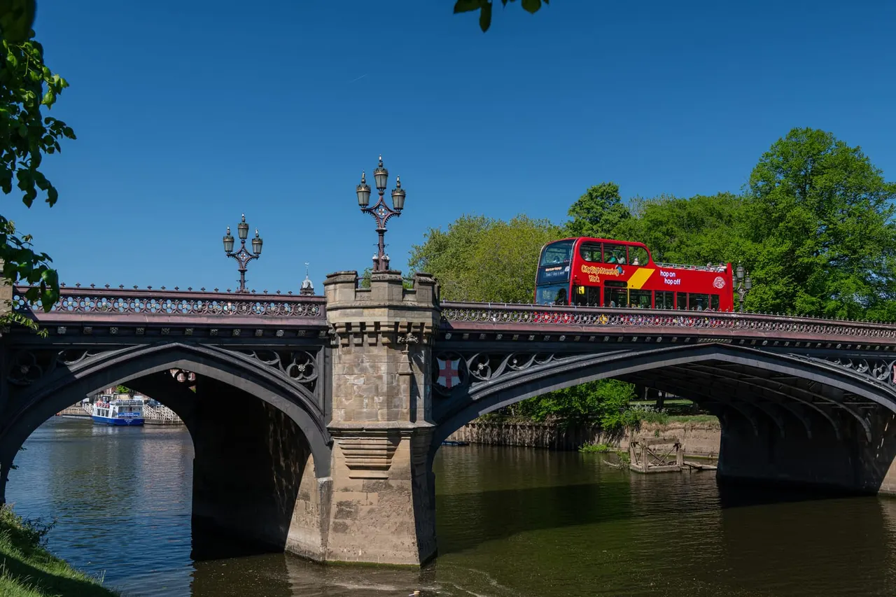 A red double-decker tour bus crosses an ornate stone bridge spanning a river under clear blue sky.