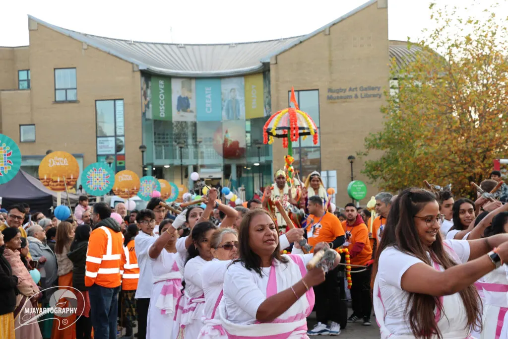 Outdoor festival celebration with large crowds wearing white and orange clothing, colourful decorative umbrellas and traditional dress parading in front of a brick building.