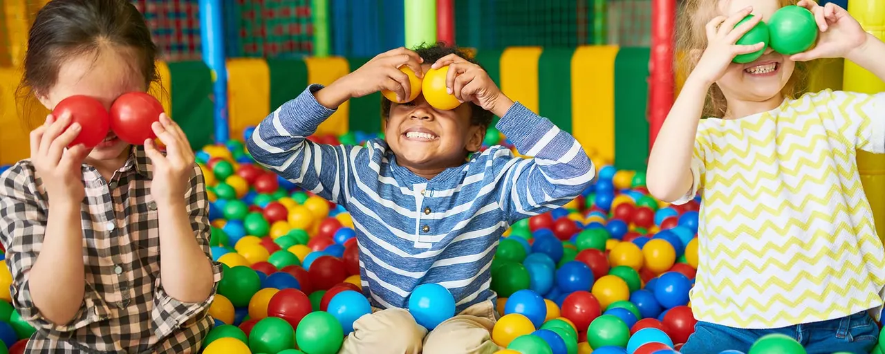 Three children playing in a ball pit filled with colourful plastic balls, holding balls up to their faces and smiling.