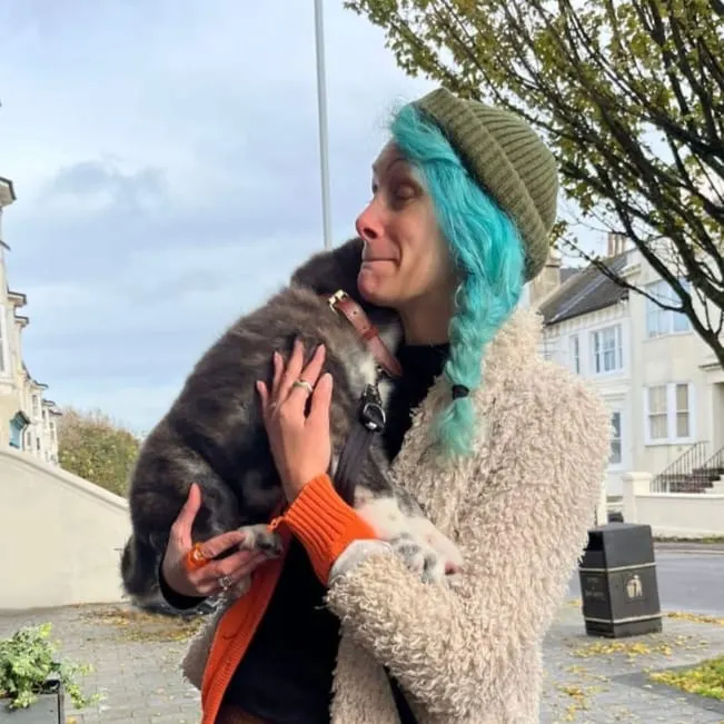 Woman with turquoise hair holding a young fox in an urban courtyard setting.