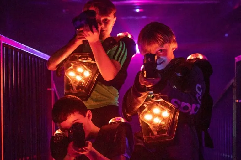 Three children holding illuminated laser tag equipment during an indoor gaming session with purple ambient lighting.