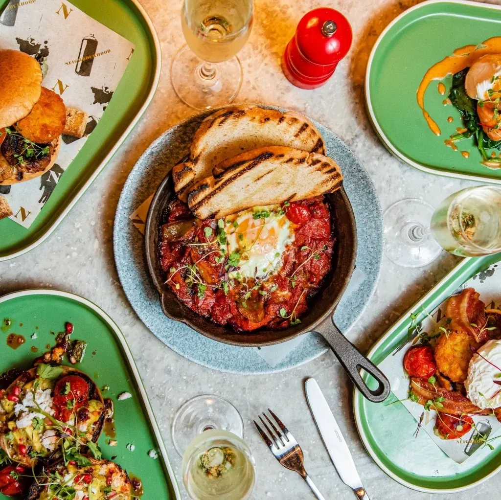Overhead view of a sharing meal with grilled meat sandwich, charred vegetables on green plates, and white wine glasses on a light table.