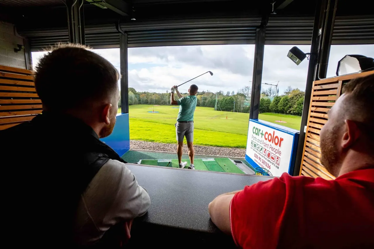 Two people watch from indoors as a golfer swings at an indoor golf driving range with a green field visible beyond.