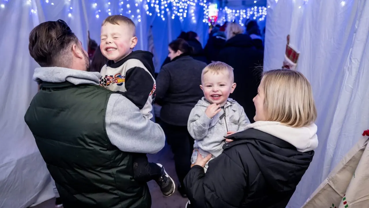 Adults and young children smiling together in an illuminated tunnel with blue and white fairy lights overhead.