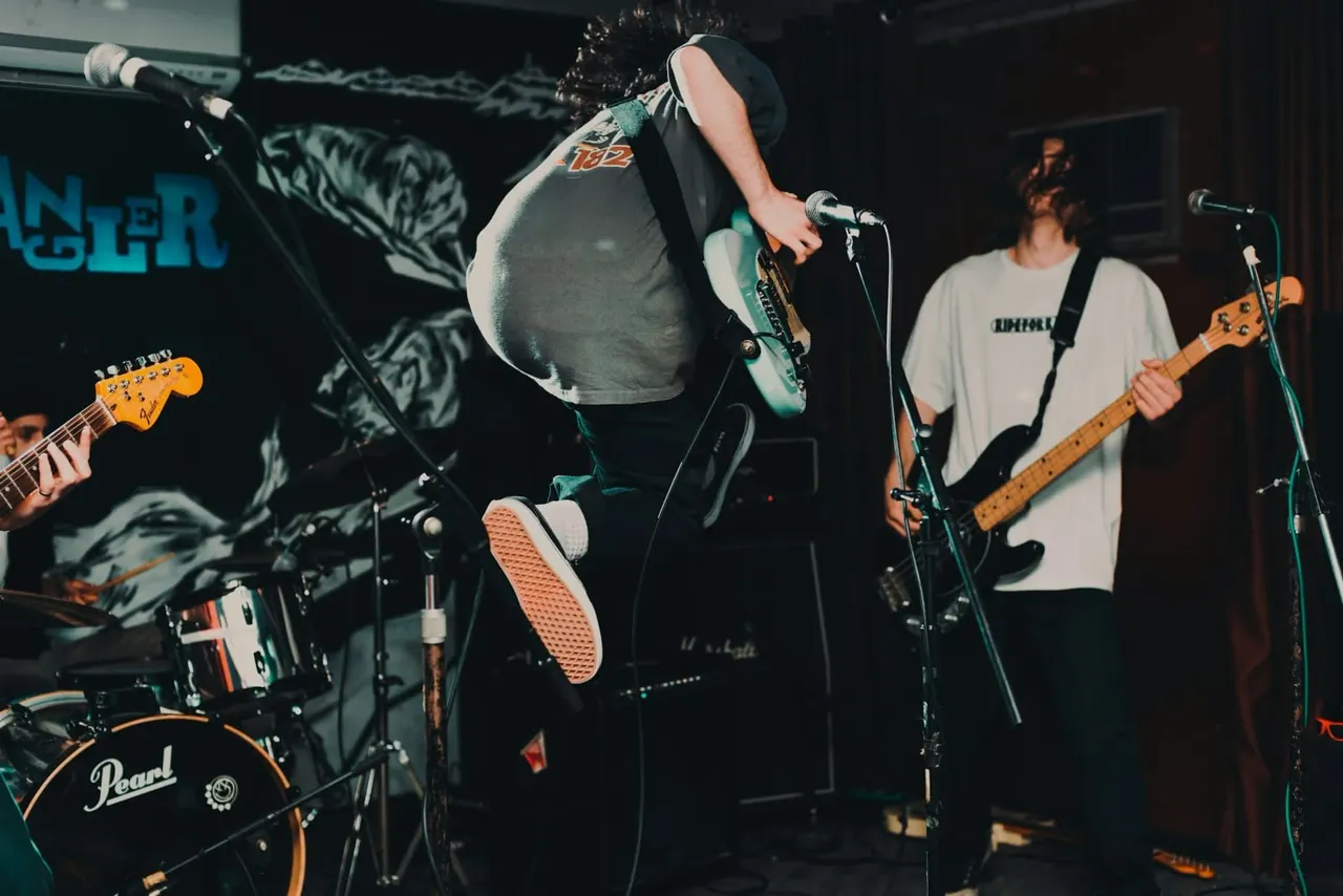 A rock band performs on stage with singer, two guitarists and drummer under stage lighting.