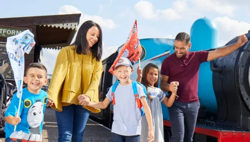 A family of two adults and three children posing happily beside a heritage railway locomotive on a sunny day.