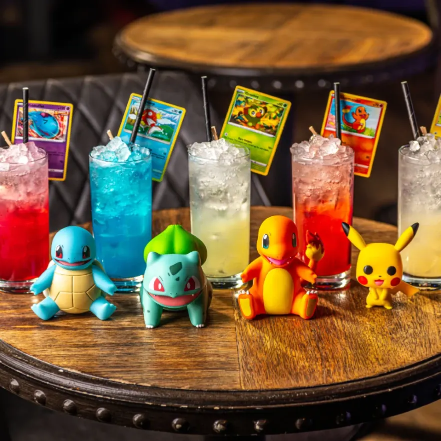 Colourful cocktails with ice and straws displayed on a wooden table alongside Pokémon character figurines and trading cards.
