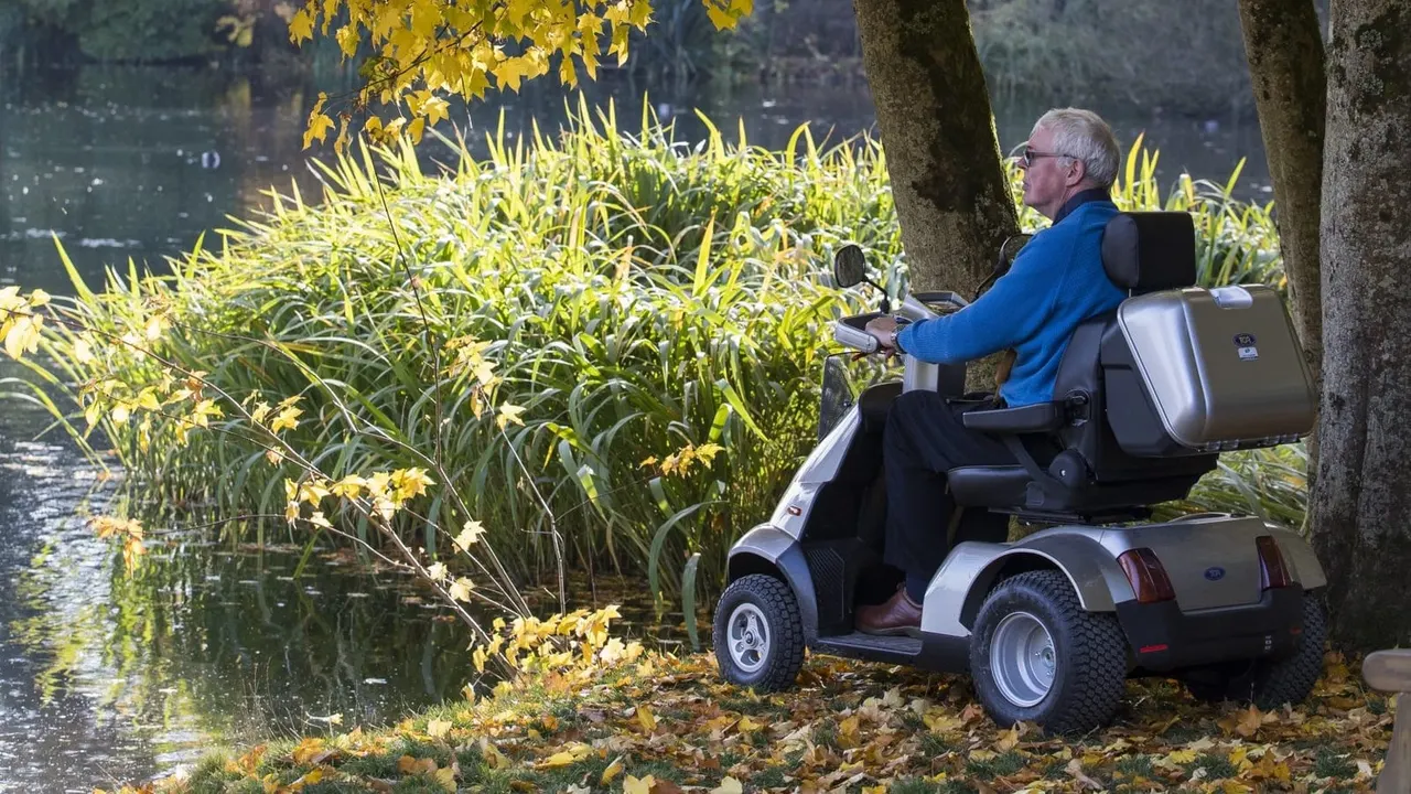 Older man on a mobility scooter fishing by a lake's edge in autumn woodland.