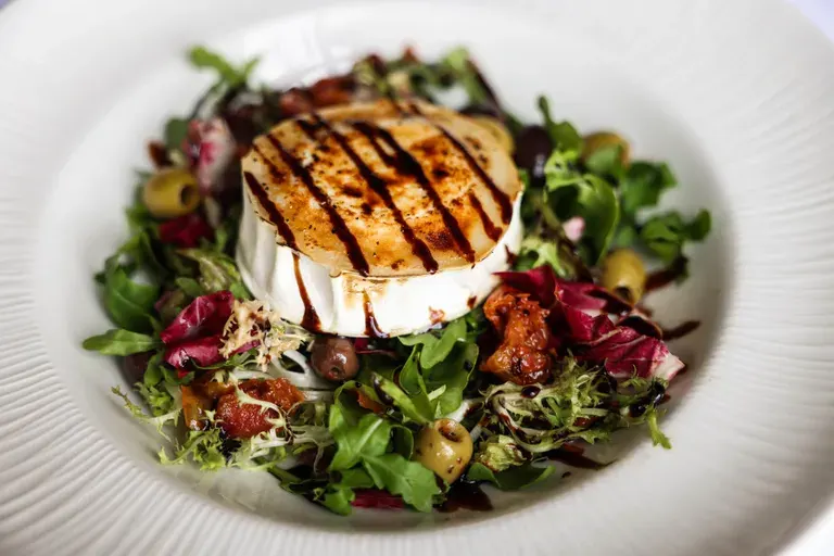 Warm goat's cheese topped with honey drizzle served on a bed of mixed greens and roasted vegetables on a white plate.