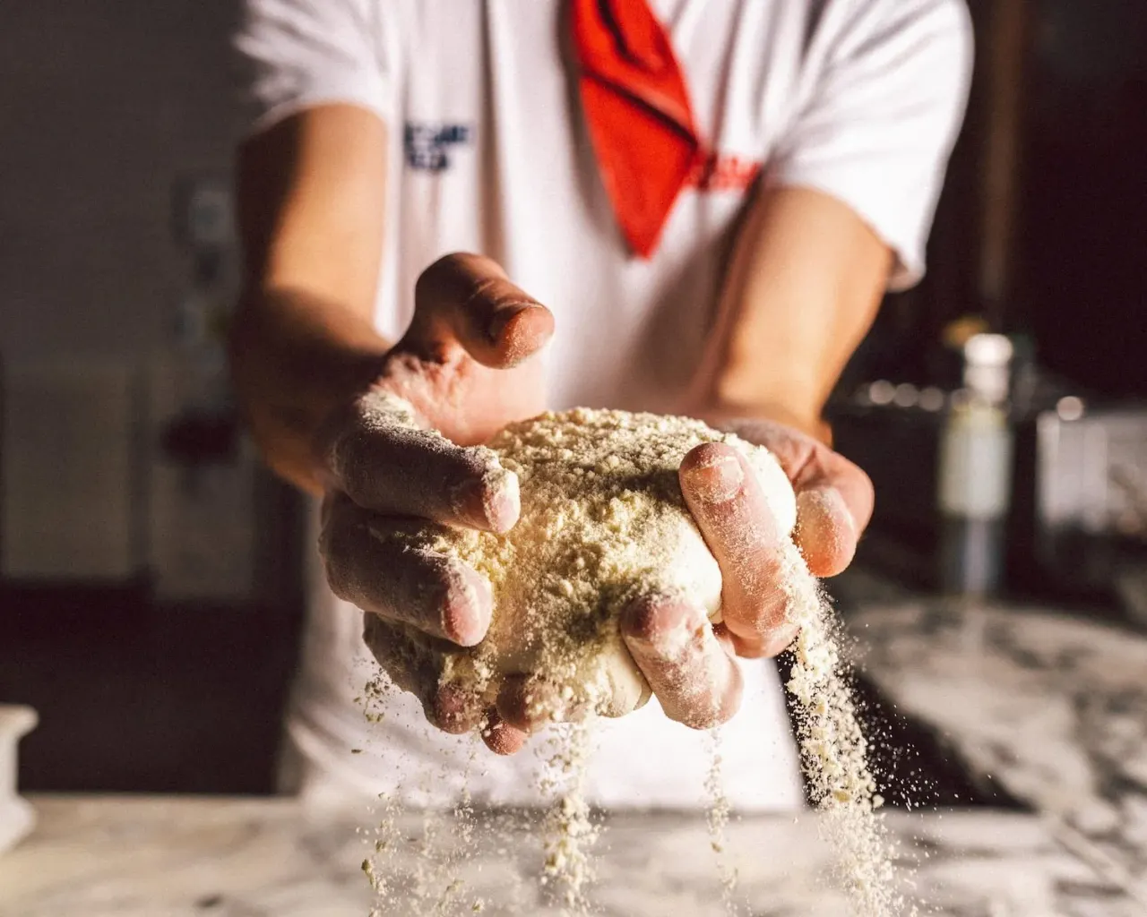 Baker in white shirt and red neckerchief holds handful of flour, letting it fall through fingers onto work surface.
