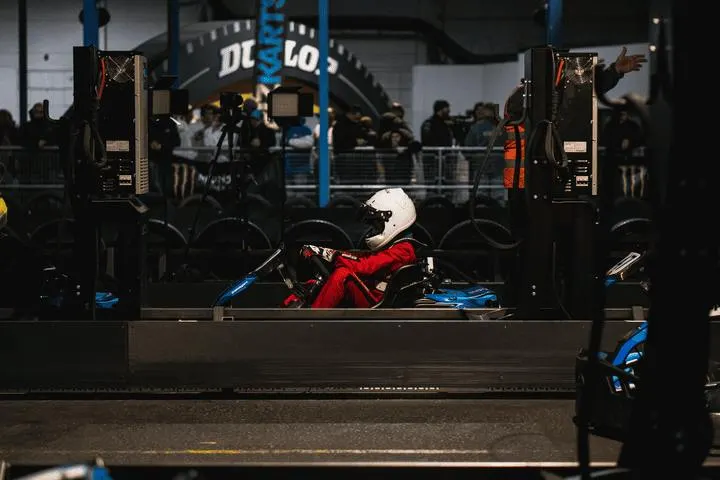 Child in white helmet and red suit sits in blue go-kart at indoor racing track with spectators behind barrier.