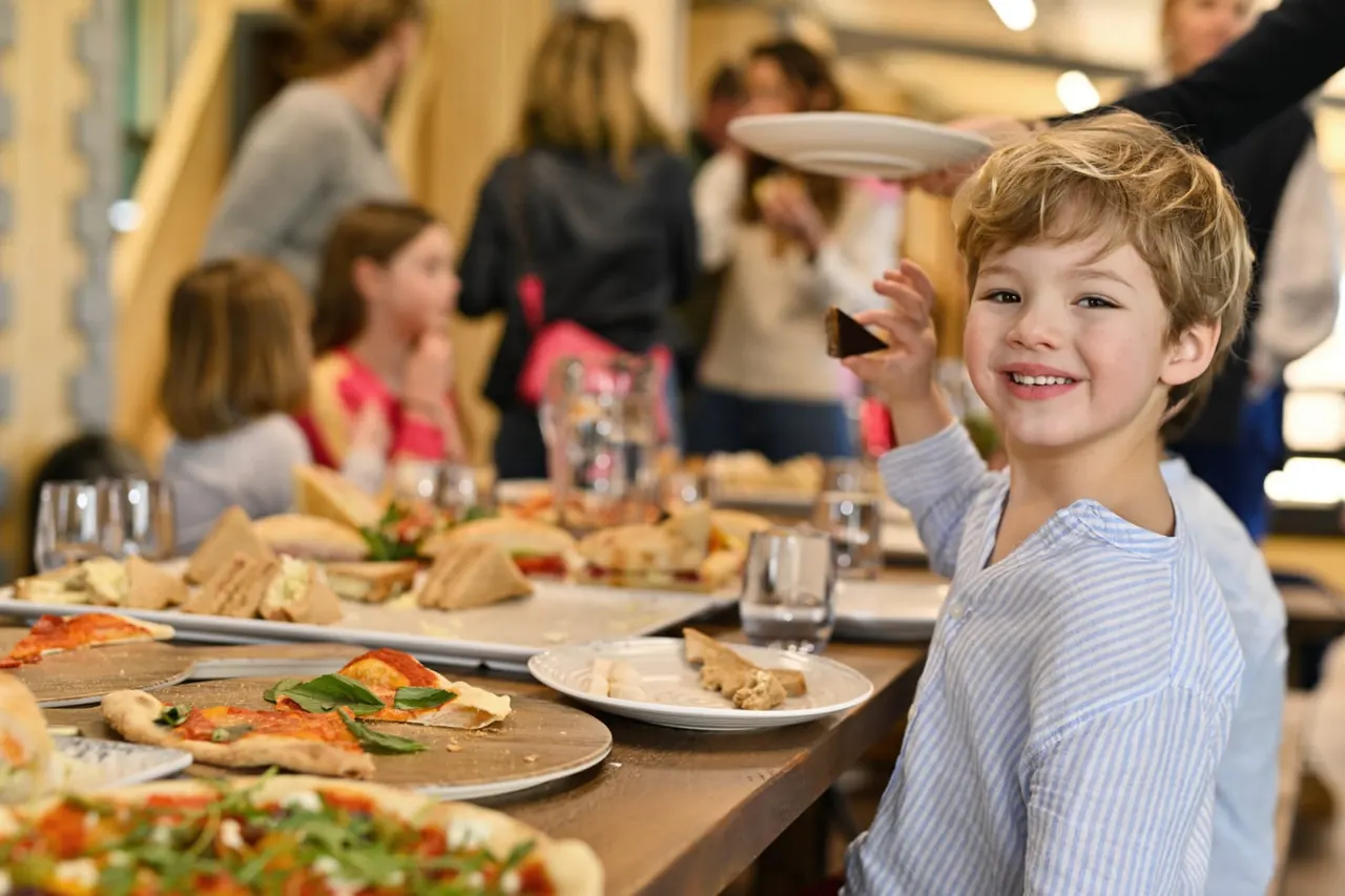 A young boy in a blue striped shirt smiles at a dining table with pizza and other food, whilst other children and adults eat and socialise behind him.