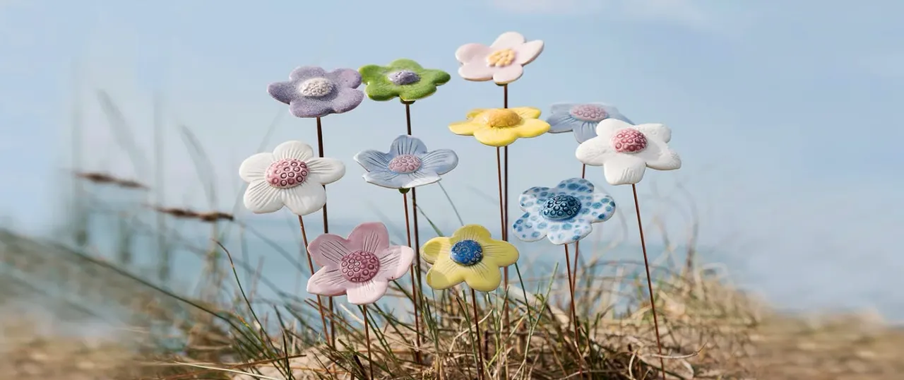 Colourful ceramic flowers with patterned centres displayed on a sandy beach with grass and a building in the background.