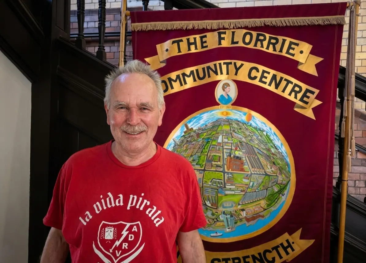 Older man with grey hair and moustache wearing a red t-shirt stands smiling in front of The Florrie Community Centre banner.
