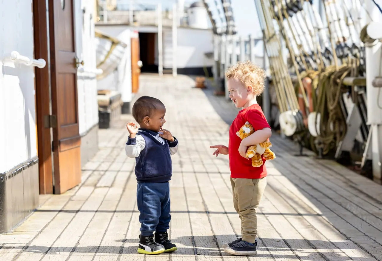 Two young children play together on the deck of a moored sailing vessel, surrounded by ropes and rigging.