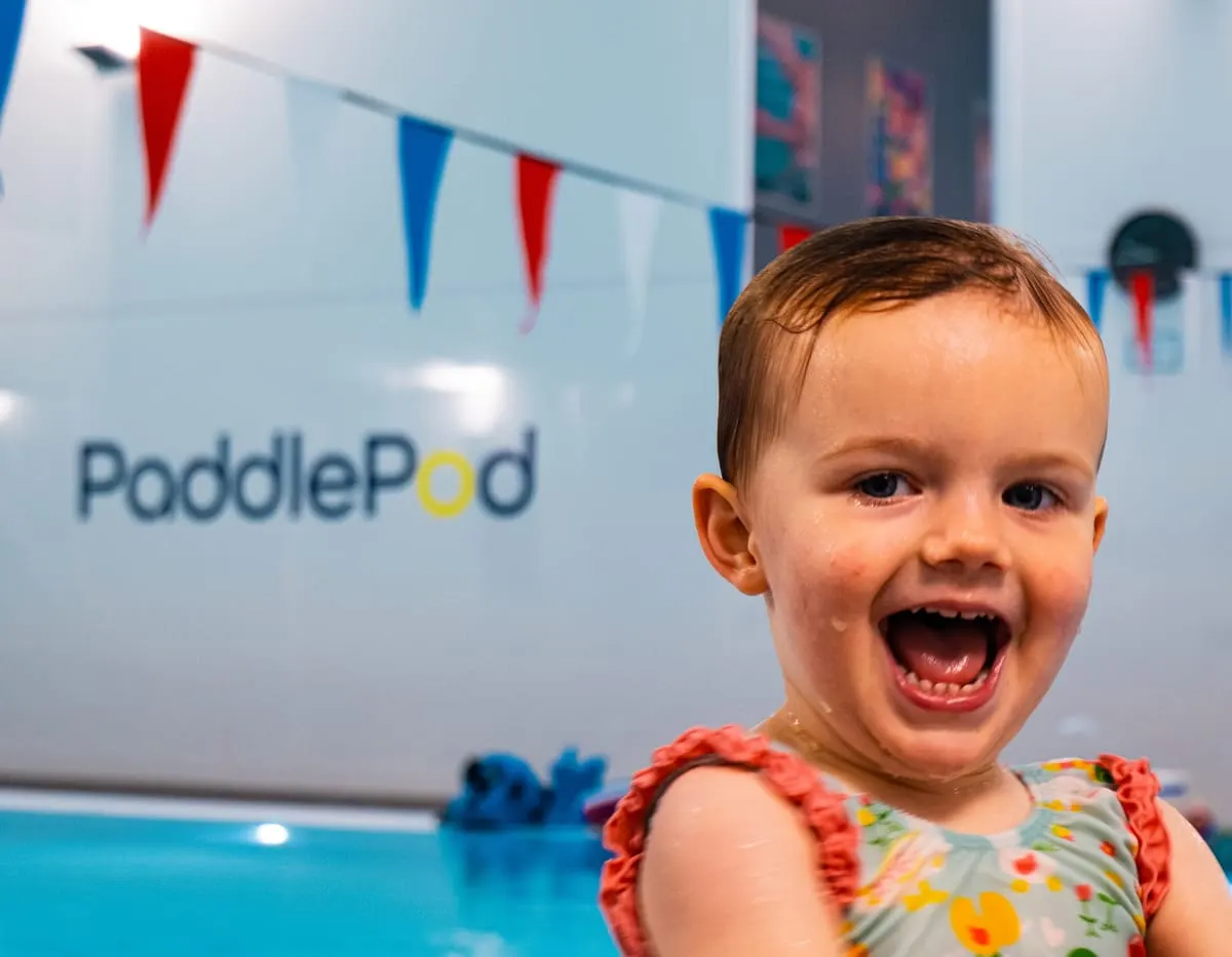 Smiling toddler in colourful swimwear at an indoor pool with bunting and PaddlePod signage.