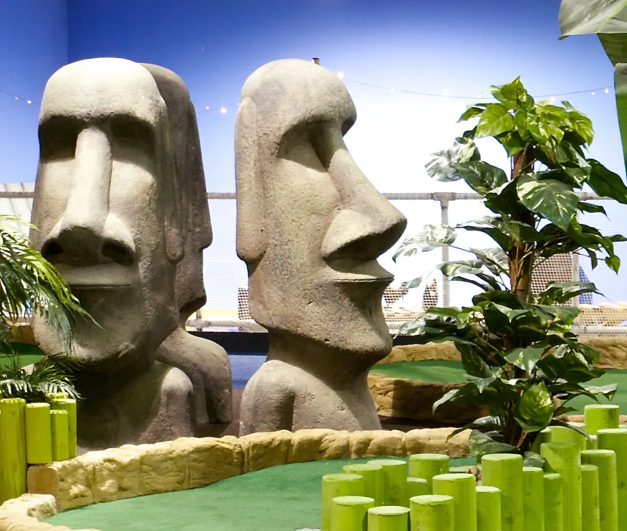 Stone moai statues flanking a minigolf hole with tropical plants and bridge backdrop under clear blue sky.