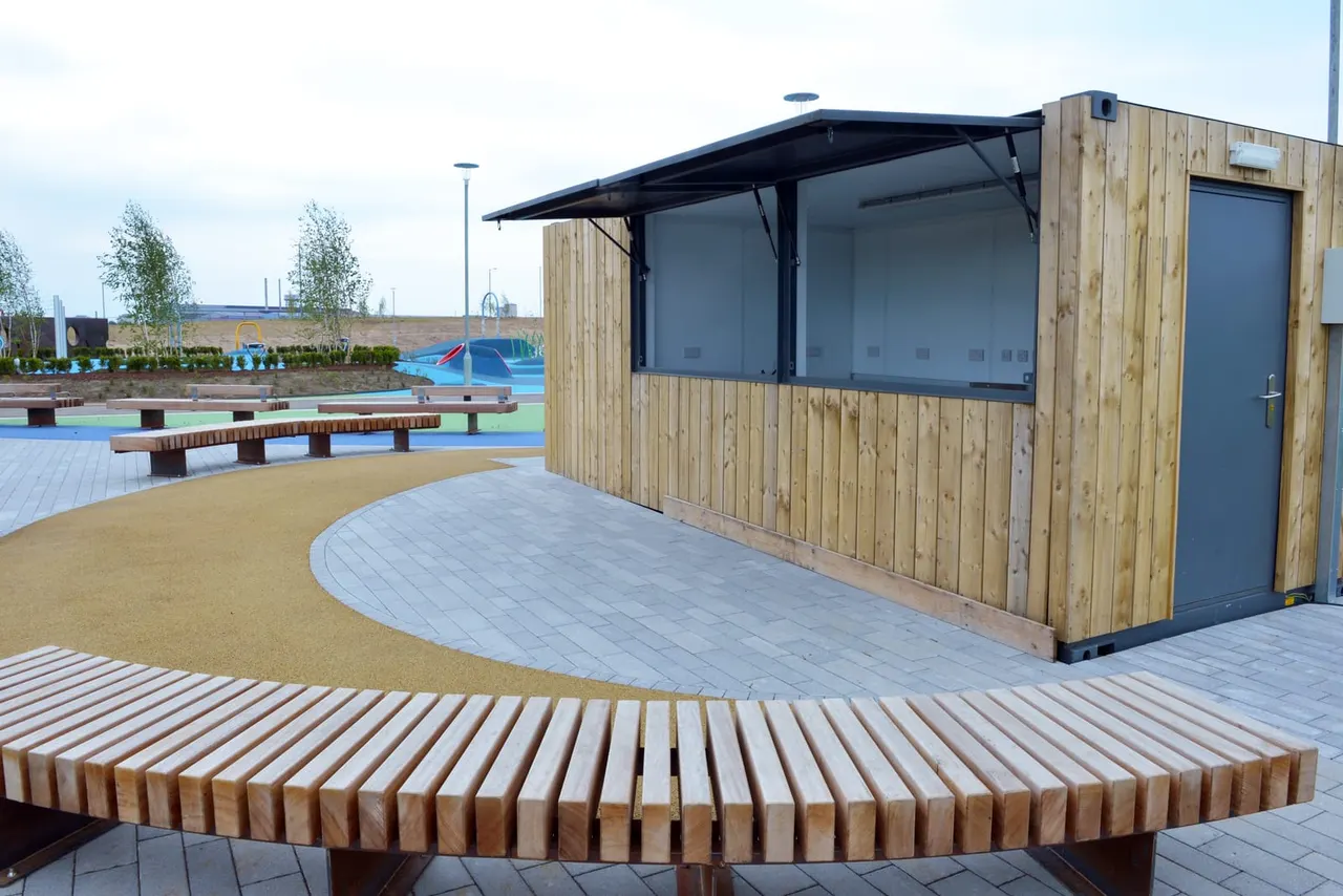 Modern wooden kiosk with service counter overlooking a play park with coloured play equipment and wooden seating areas.