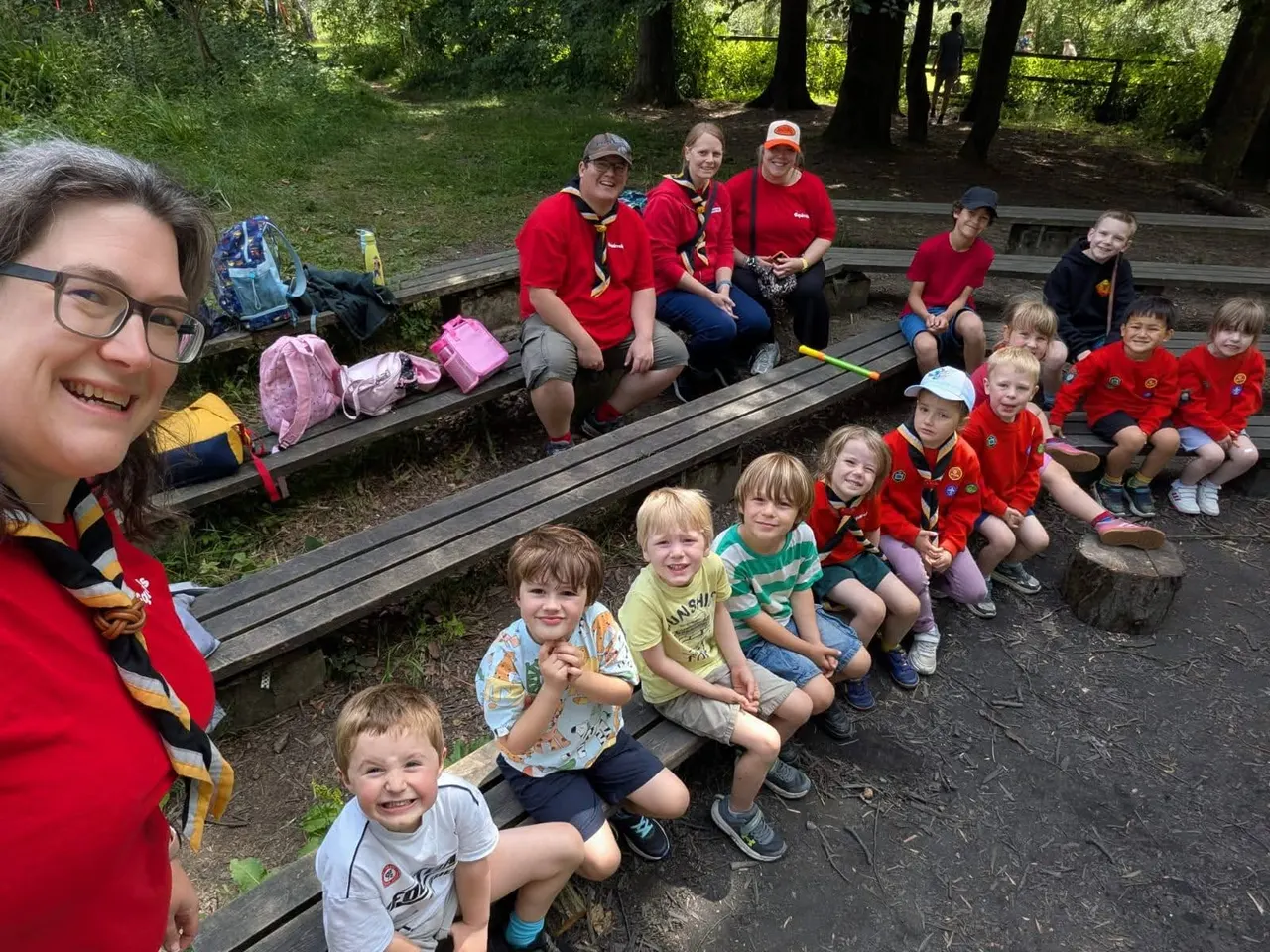 Scout leaders and children gathered on wooden benches in a woodland clearing during an outdoor group activity.