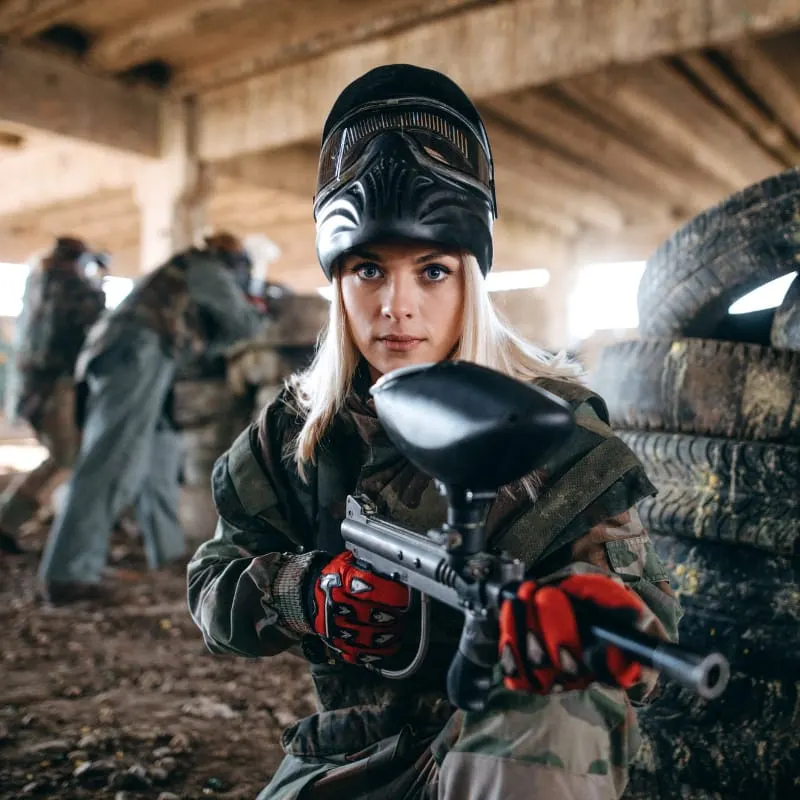 Woman in protective gear holding a paintball gun at an indoor paintball venue with other players in the background.