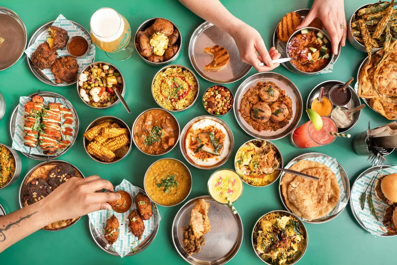 Overhead view of hands reaching for shared Indian street food dishes on a green table.