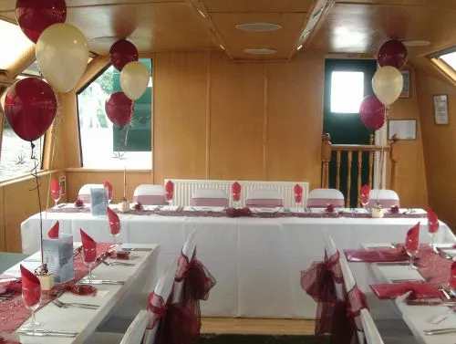 Decorated event space on a river boat with tables set for a formal occasion, burgundy and cream balloons, and place settings with napkins.