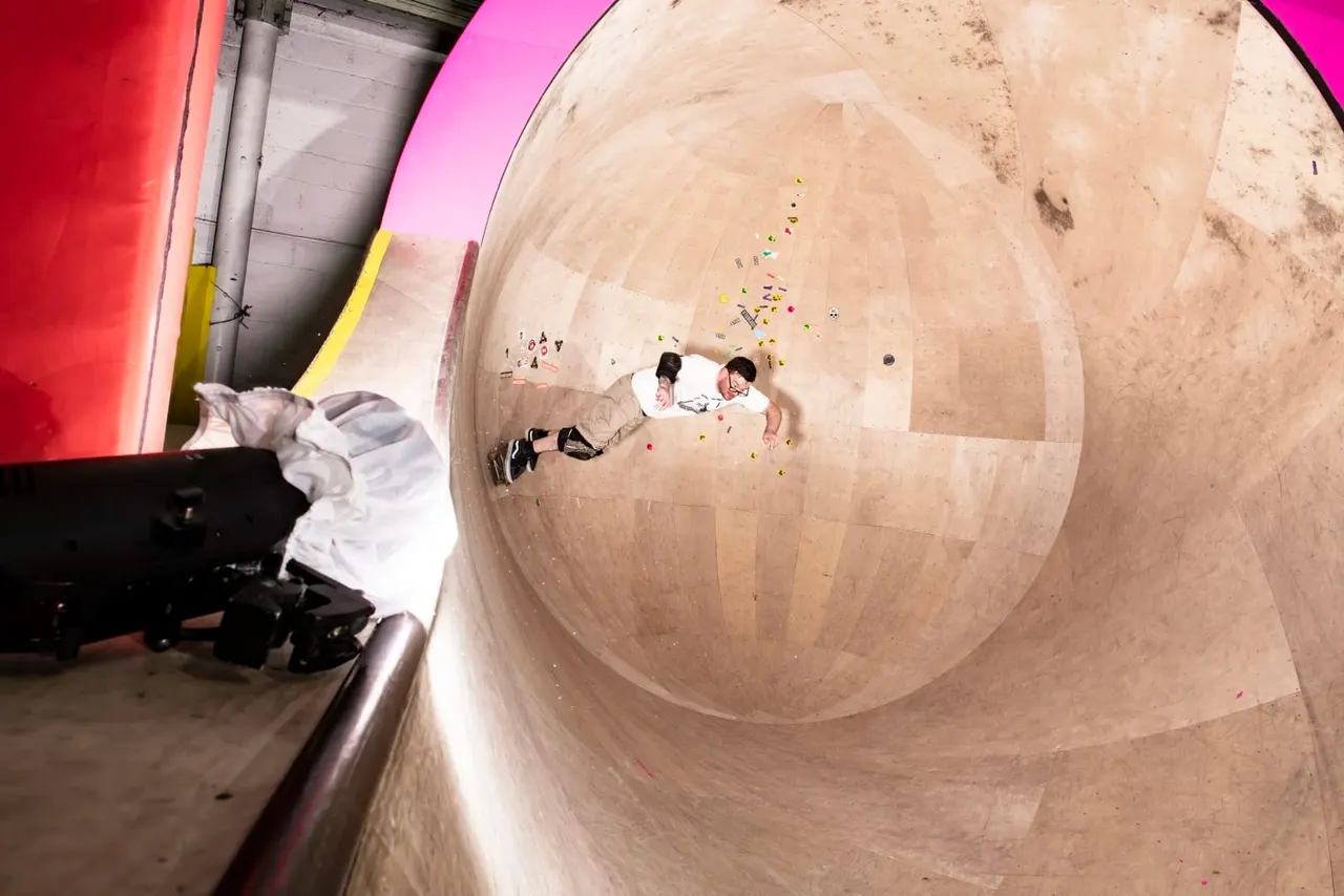 Two children sliding down the interior of a large concrete bowl-shaped skate ramp with coloured markers scattered across the surface.