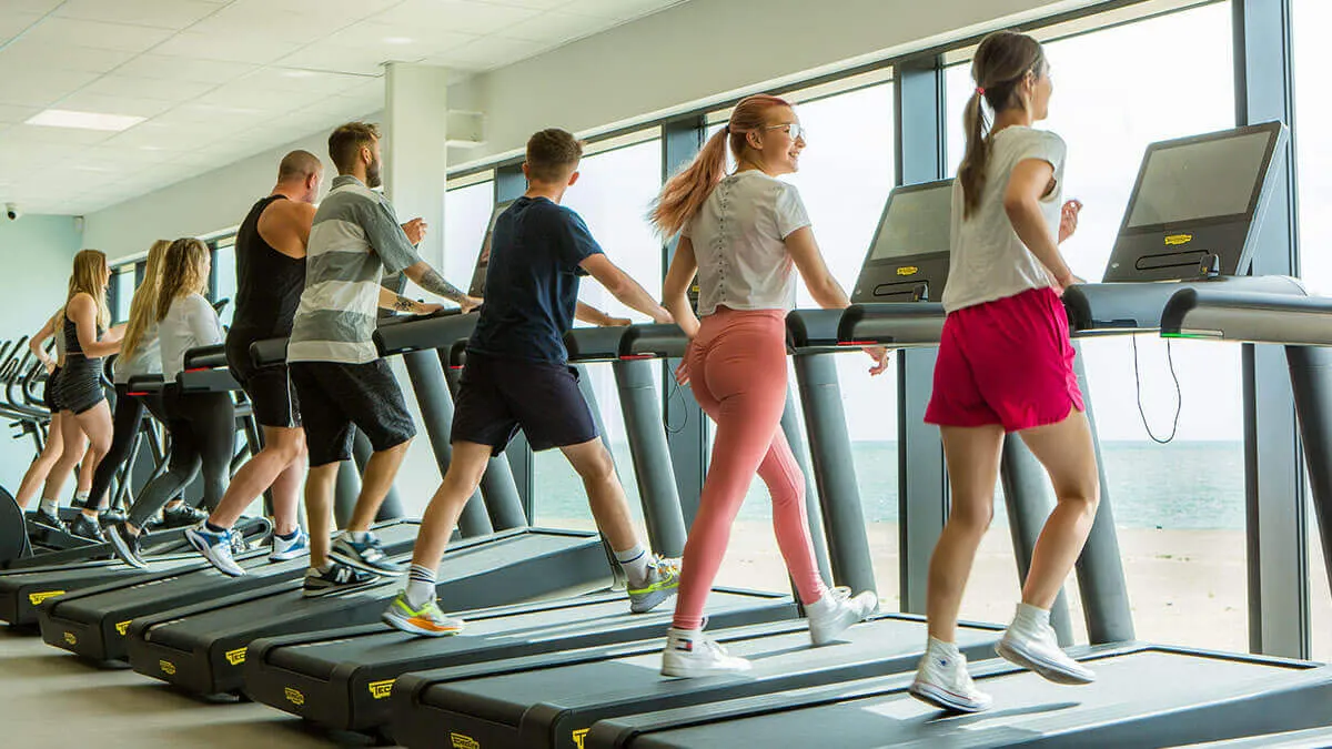 Group of people running on treadmills in a bright gym with sea views through large windows.