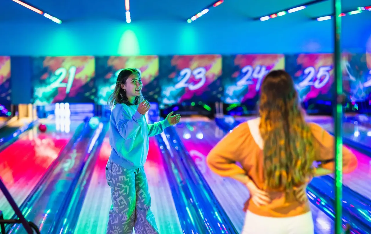 Two children bowling at an illuminated neon bowling alley with colourful lane lighting and digital scoring screens overhead.