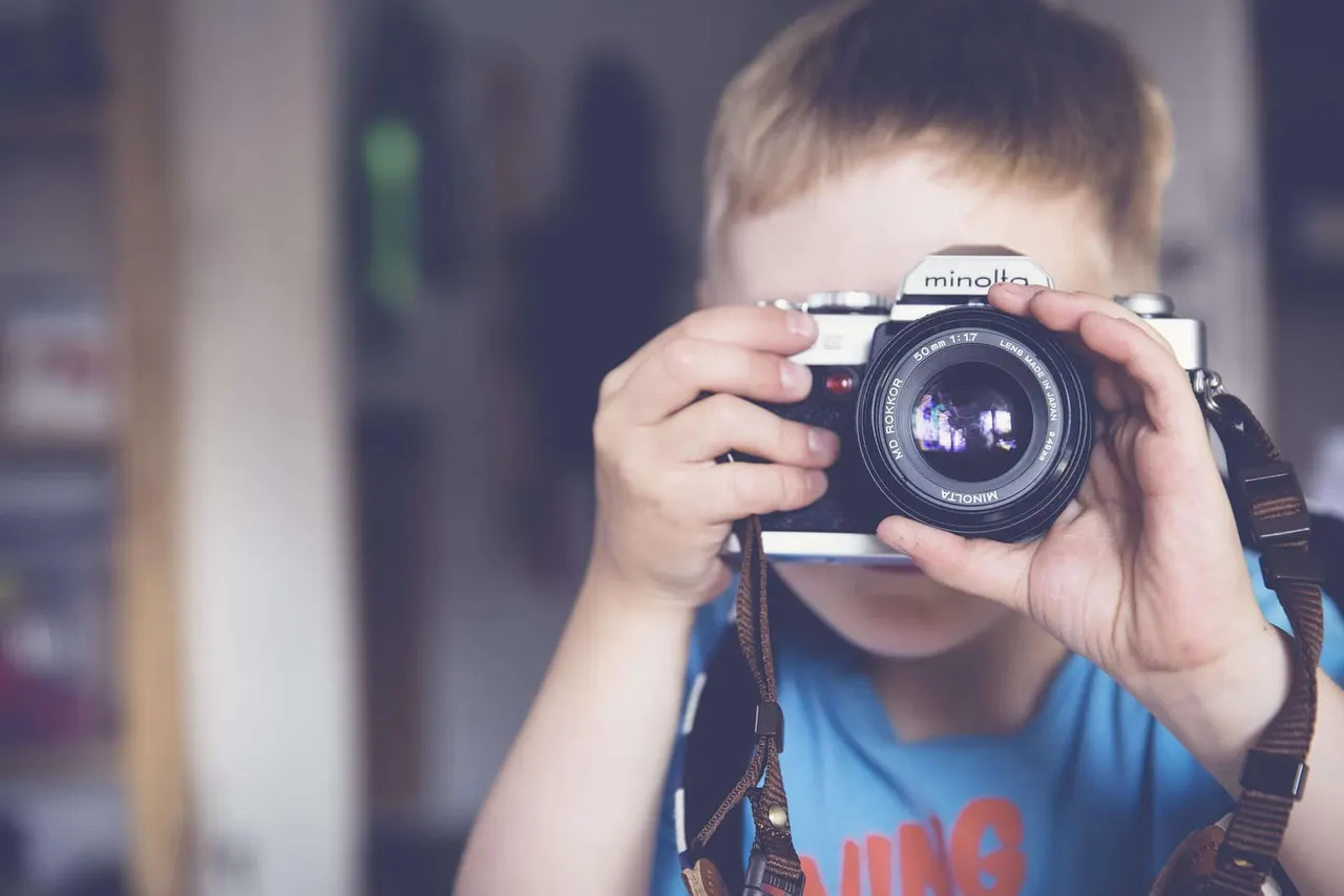 A young child in a blue shirt holds a vintage film camera up to their face, looking through the viewfinder.