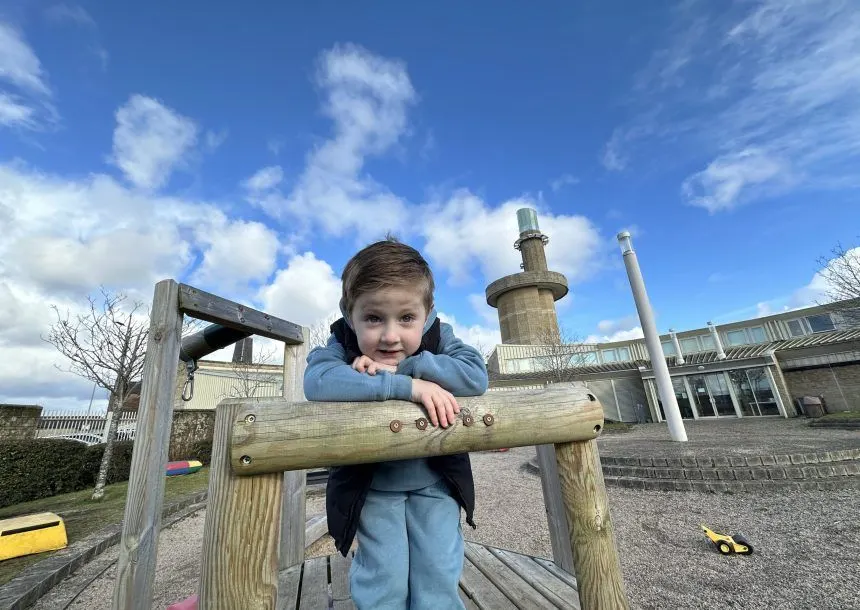 Young child smiling at wooden play equipment with heritage buildings in background under blue sky.