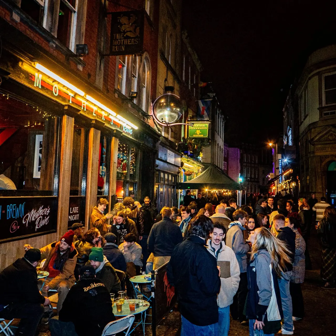 Crowded street scene at night outside a pub with people drinking and socialising at tables and standing in a narrow alleyway lined with illuminated shop fronts.