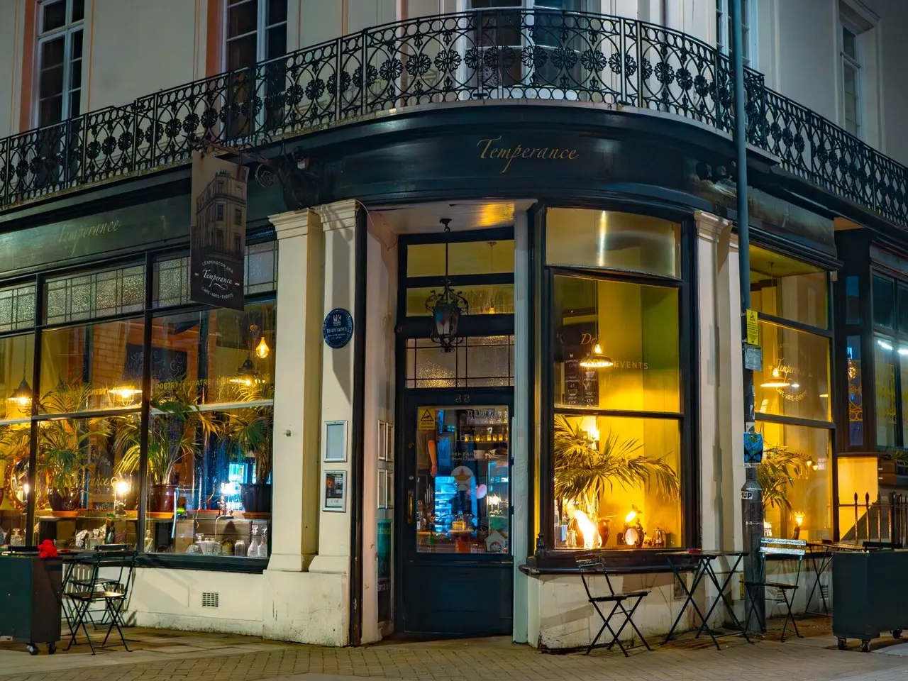 Corner bar and restaurant with illuminated windows, ornate wrought-iron balcony above, and outdoor seating at night.