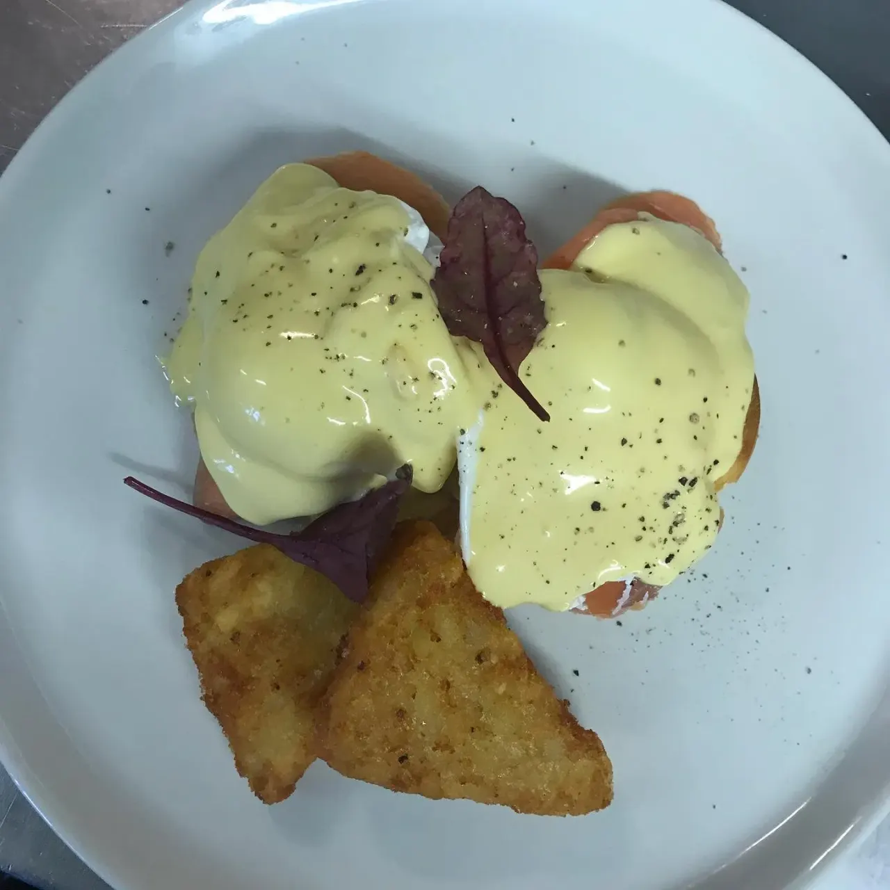 Eggs Benedict on toasted bread with hollandaise sauce, purple basil and fried potatoes on a white plate.