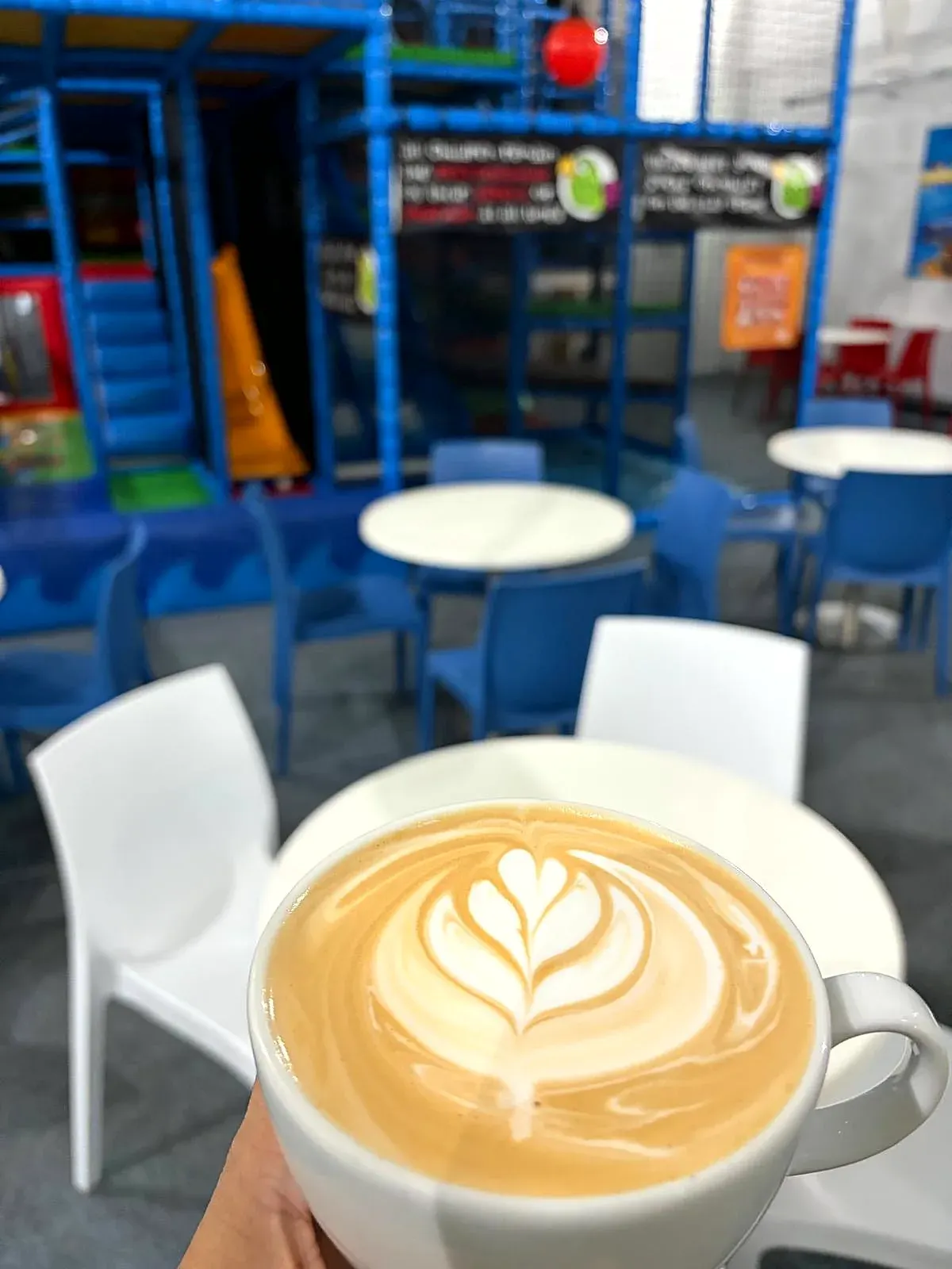 Hand holding a latte with latte art in an indoor play centre with blue furniture and climbing structures.