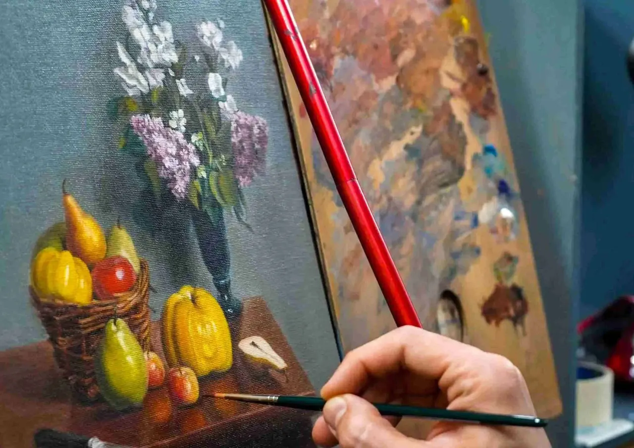 A person's hand holding a paintbrush while painting still-life artwork on canvas featuring fruit and flowers.