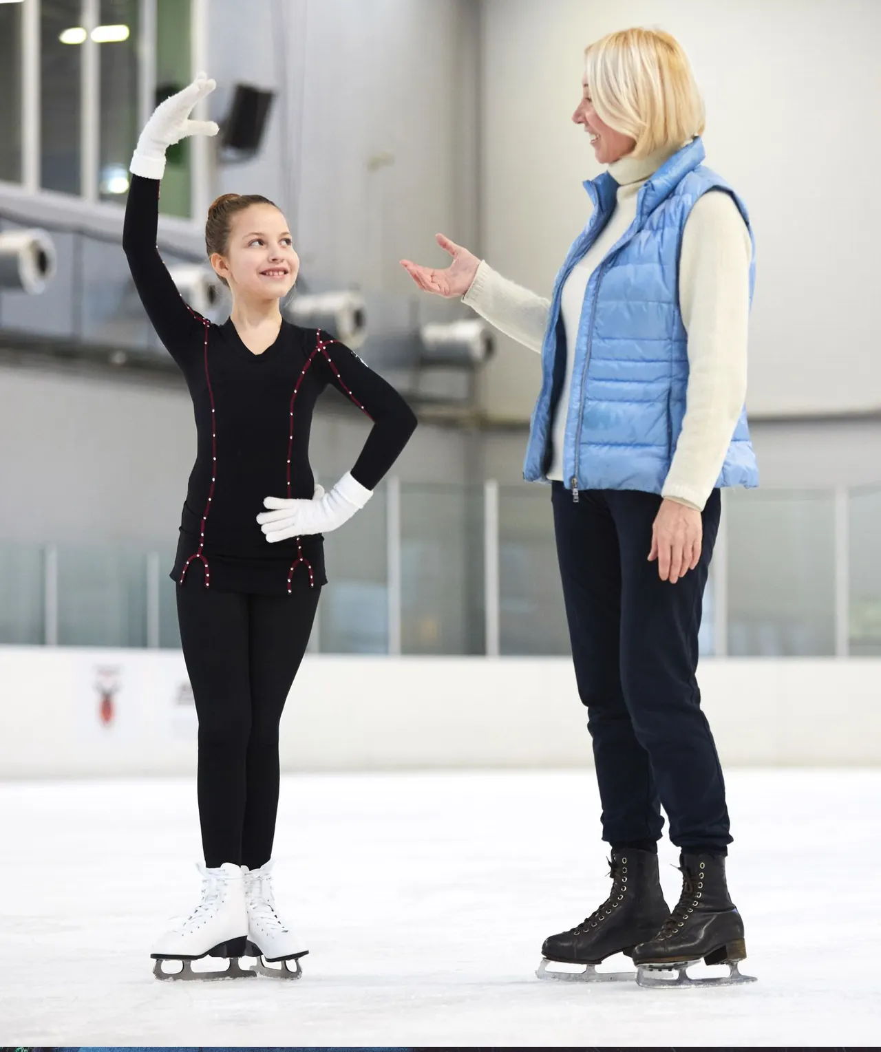 A young figure skater in black costume with red sequins poses on ice whilst an adult instructor in a blue gilet stands beside her.