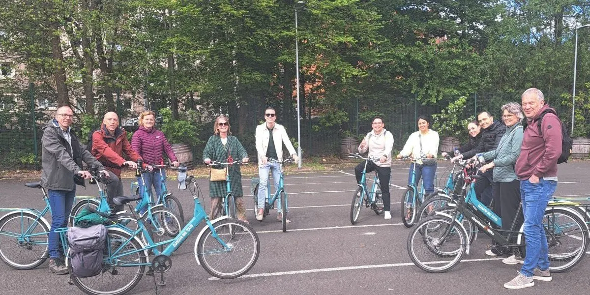 A group of adults posing with turquoise bicycles in a tree-lined park setting on a paved path.