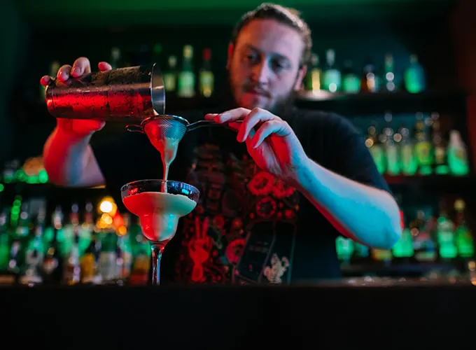 Bartender pouring a red cocktail into a glass at a bar with illuminated bottles in the background.