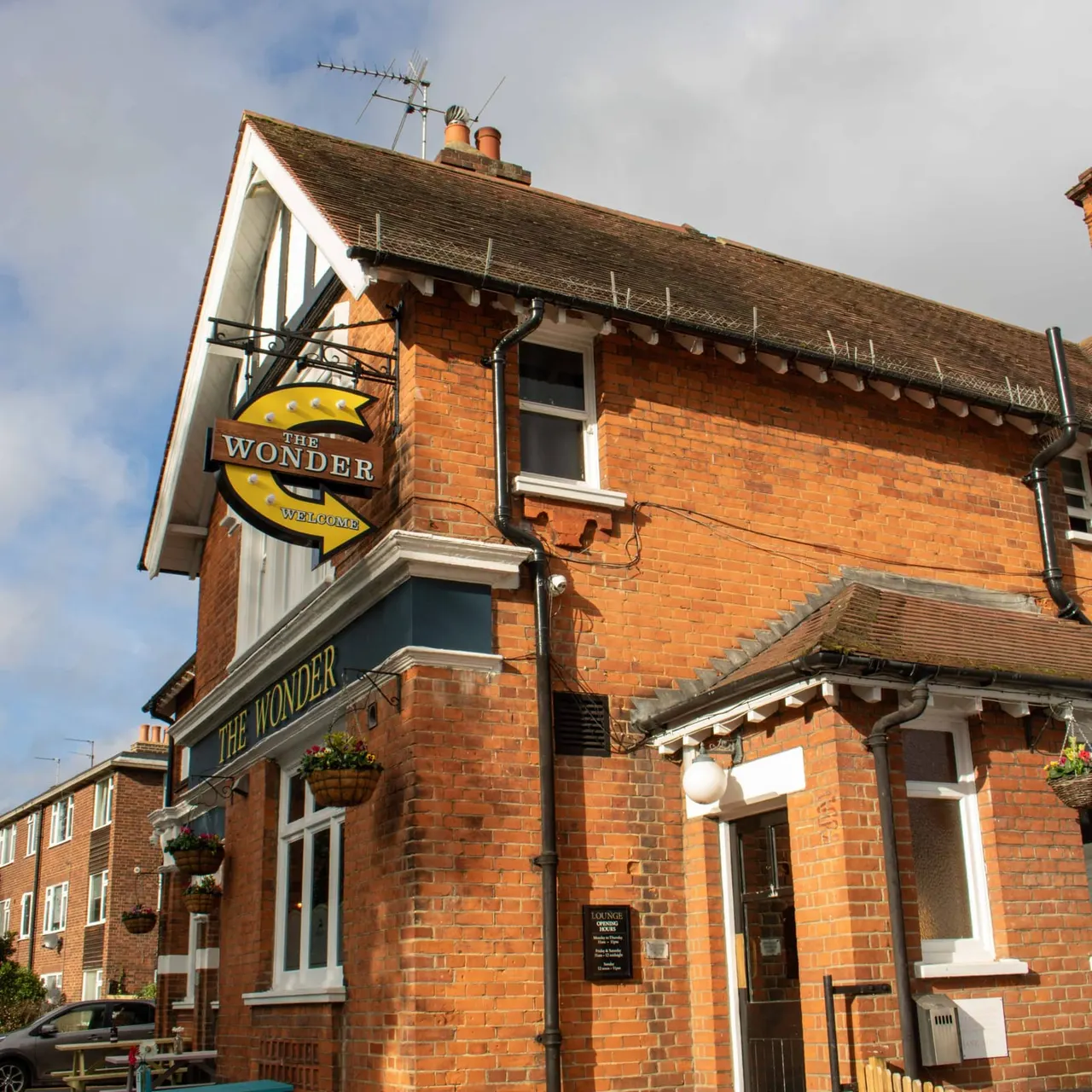 Victorian red-brick pub with white half-timbered gable, hanging baskets, and yellow "The Wonder" welcome sign.