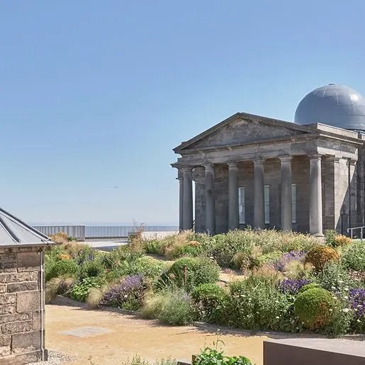Classical stone building with dome and columns surrounded by drought-tolerant plants and grasses on a hilltop.