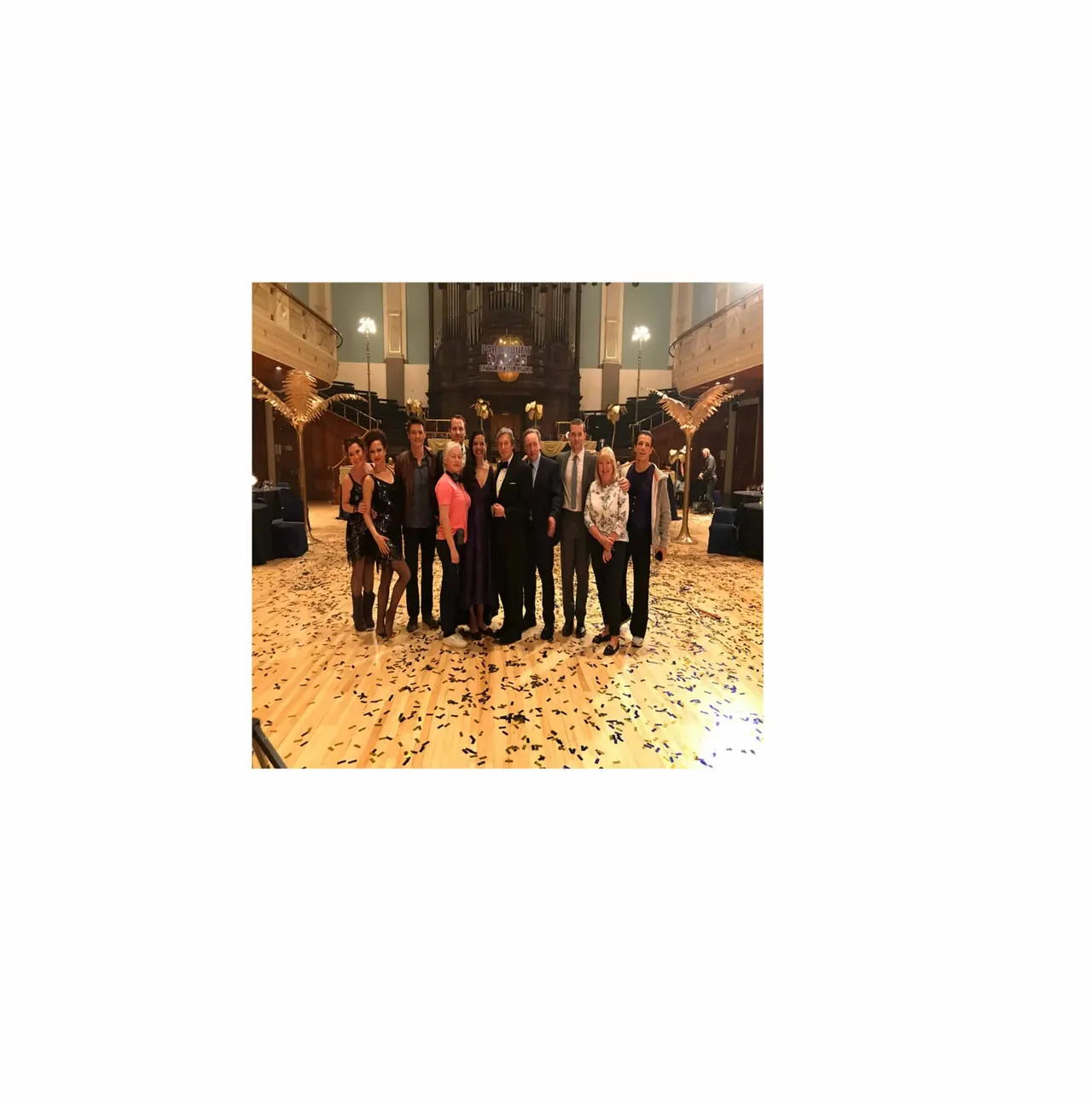 Group of formally dressed people posing together inside a grand concert hall with an organ and confetti on the floor.