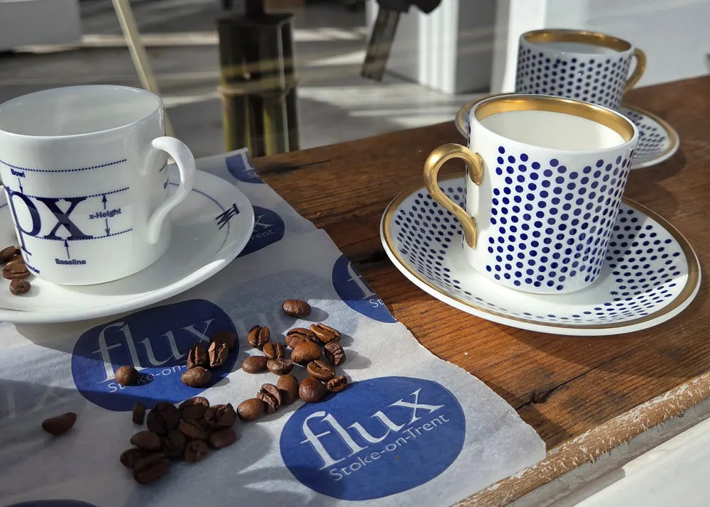 Two cups of coffee with blue and white patterned cups and saucers on a wooden table with coffee beans scattered on branded napkins.